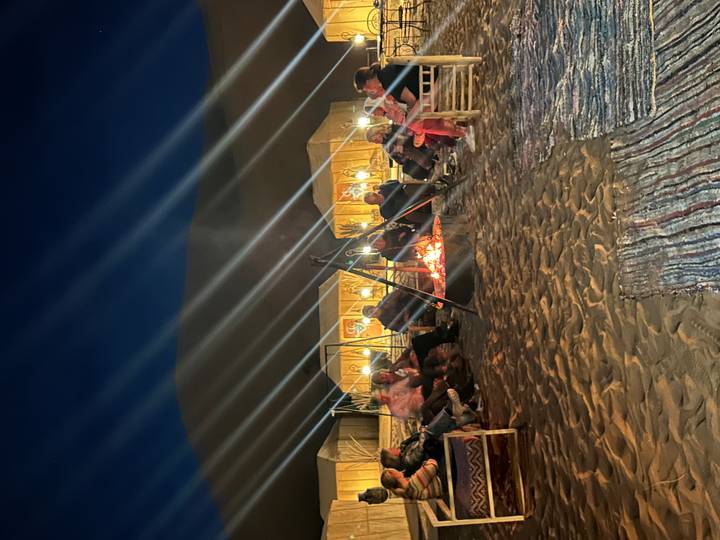 Nighttime campfire circle on desert sand with tents behind and beams of light streaking overhead.