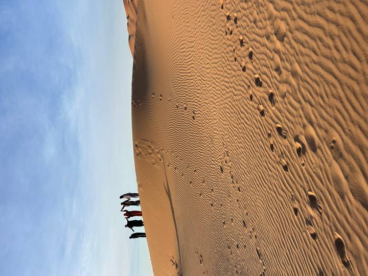Sweeping golden sand dune under a clear blue sky with a line of travelers holding hands along the ridge.
