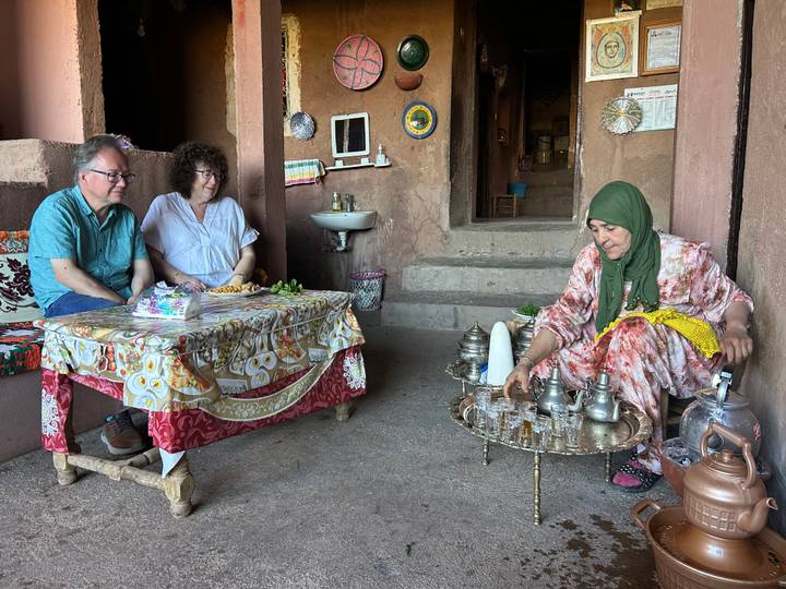 Local hostess pours traditional Moroccan mint tea for two visitors seated in a rustic adobe home.