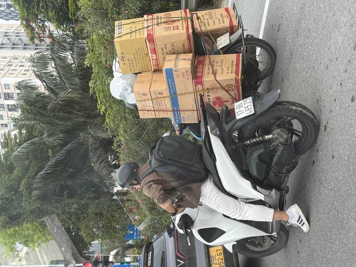 Motorbike rider with helmet balances stacks of cardboard boxes tied to the back in busy city traffic.