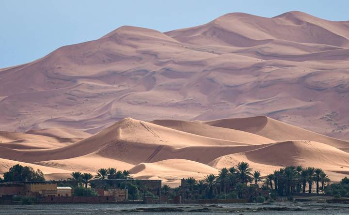 Sweeping pink desert dunes tower above a line of palms and small buildings.