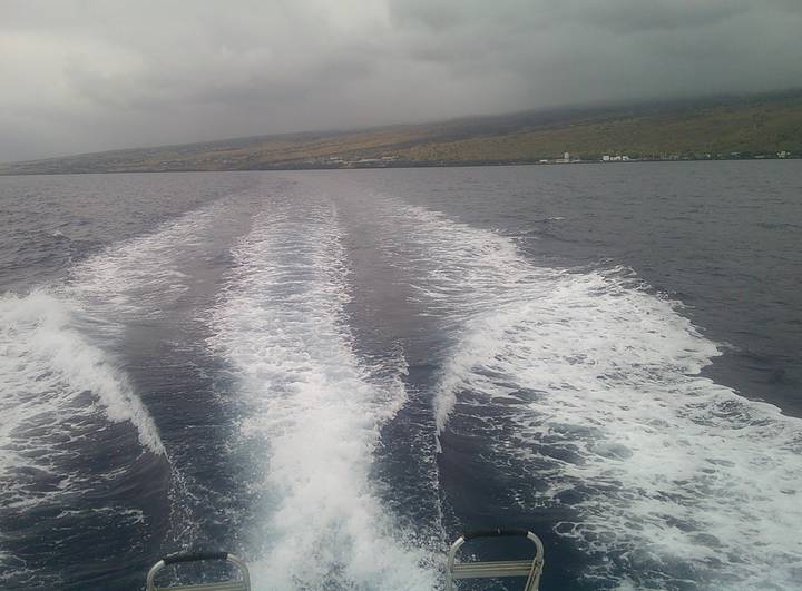 Boat wake trails across deep blue ocean with distant volcanic coastline under grey sky