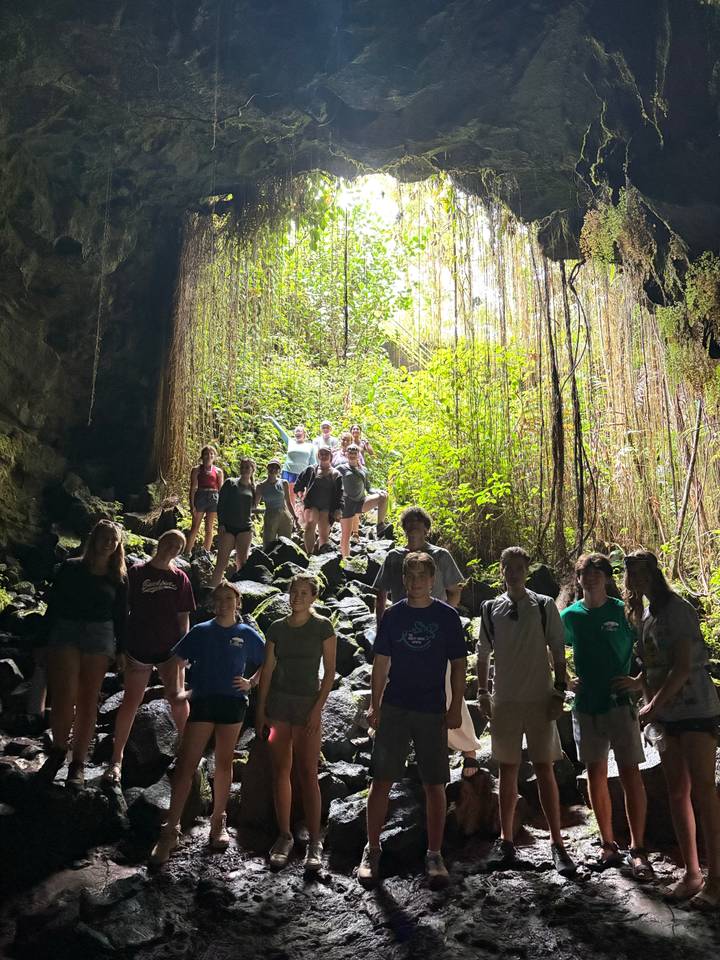 Tour group poses inside the mouth of a lush lava tube cave, strong backlighting from entrance.