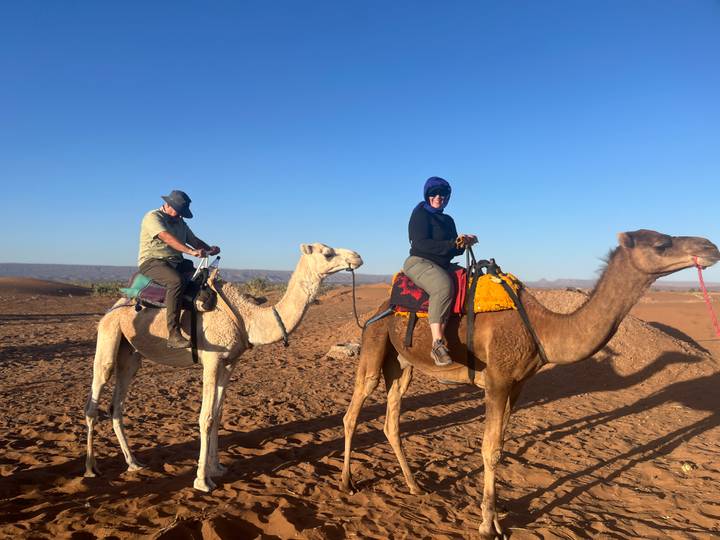 Two travellers ride camels across reddish desert sand under a vivid blue sky.