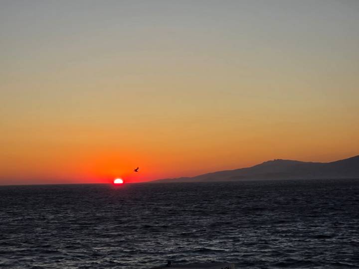 Red sun touches the Aegean horizon with orange gradient sky and distant hills.