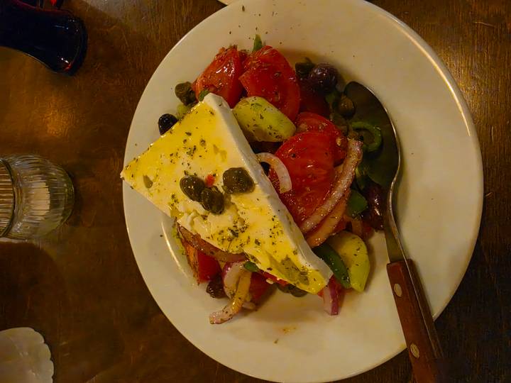 Plate of Greek salad topped with a thick feta slice, olives and herbs.