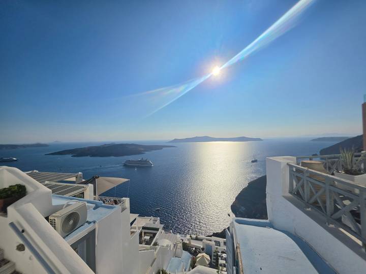 High noon sun glints over caldera waters with cruise ships anchored beside white cliff villages.