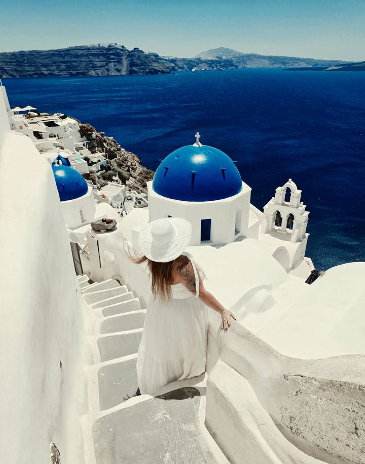 Traveller in white dress admires iconic blue domes and bell tower above deep blue Aegean.