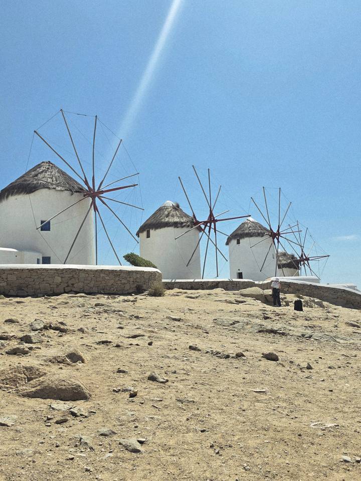 Iconic line of white windmills with red sails stand on rocky Mykonos hillside under clear sky.