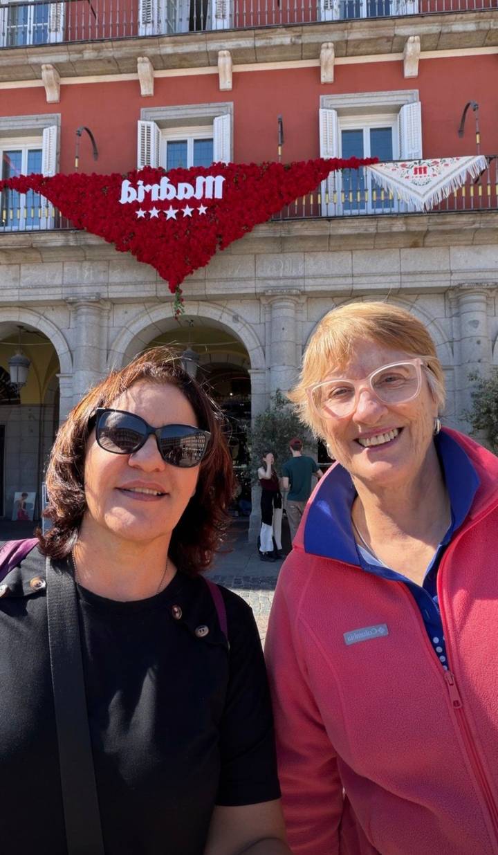 Two women smile for a selfie in front of a grand stone arcade in a sunny plaza.