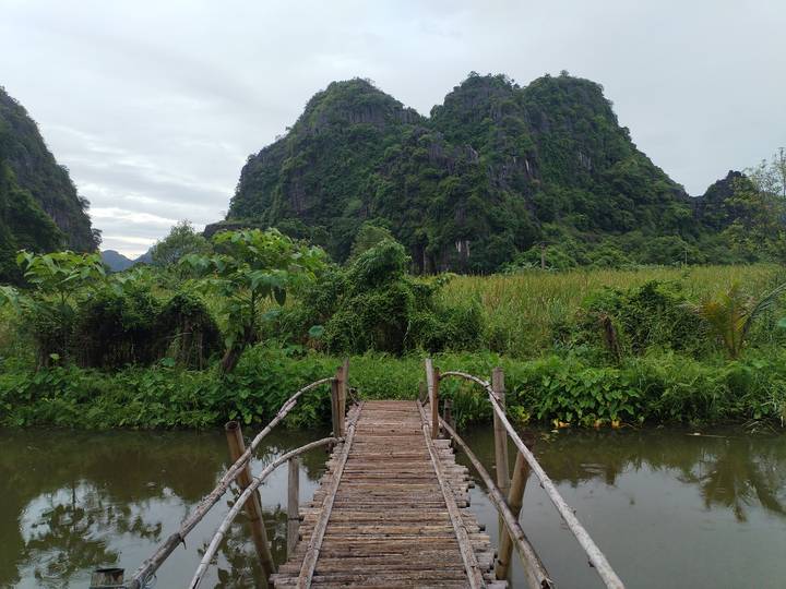 A rustic wooden footbridge crosses a marshy stream toward steep green karst hills.