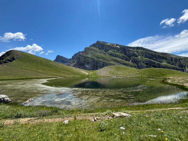 A crystal-clear alpine lake mirrors jagged green mountains under a bright blue sky.