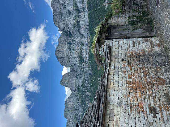 Stone wall foreground leads to towering limestone cliffs under a deep blue sky with small clouds.