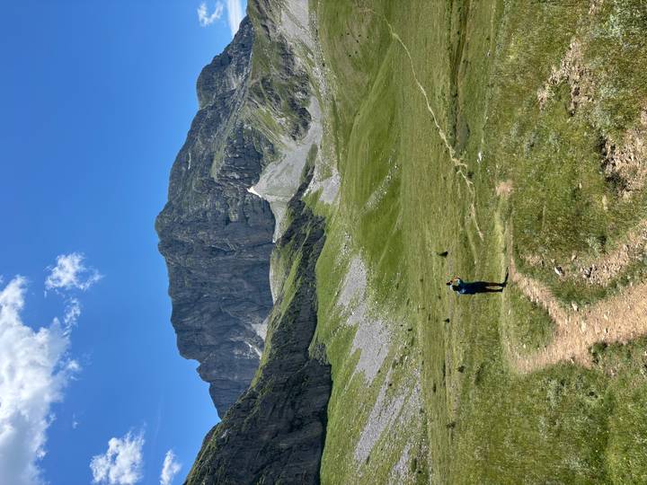 A lone hiker follows a winding trail across a green alpine valley toward a sheer mountain face.