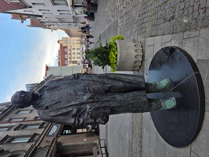 Bronze statue of a man in a long coat stands on a pedestrian street lined with historic buildings.