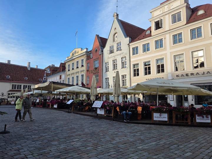 Outdoor cafés with umbrellas fill a medieval town square lined with pastel gabled buildings.