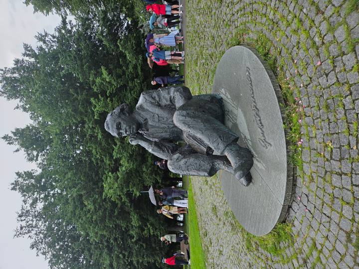 Bronze seated statue of a man rests on a circular plinth in a leafy park as visitors look on with umbrellas.