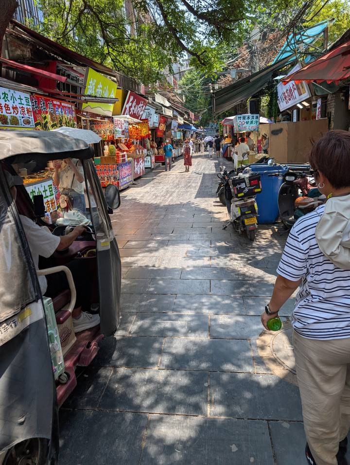 Bustling pedestrian alley lined with street food stalls, scooters and a passing tuk-tuk.