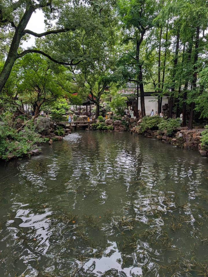 Lush classical Chinese garden with visitors walking bridges around a tranquil reflective pond.