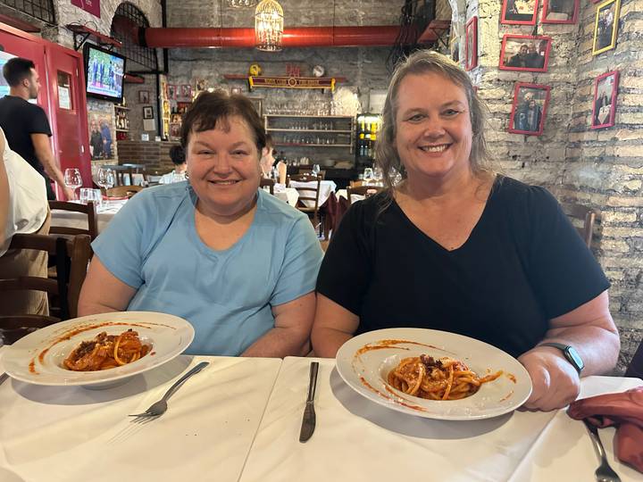 Two women smiling in a cozy brick-walled restaurant holding plates of pasta.