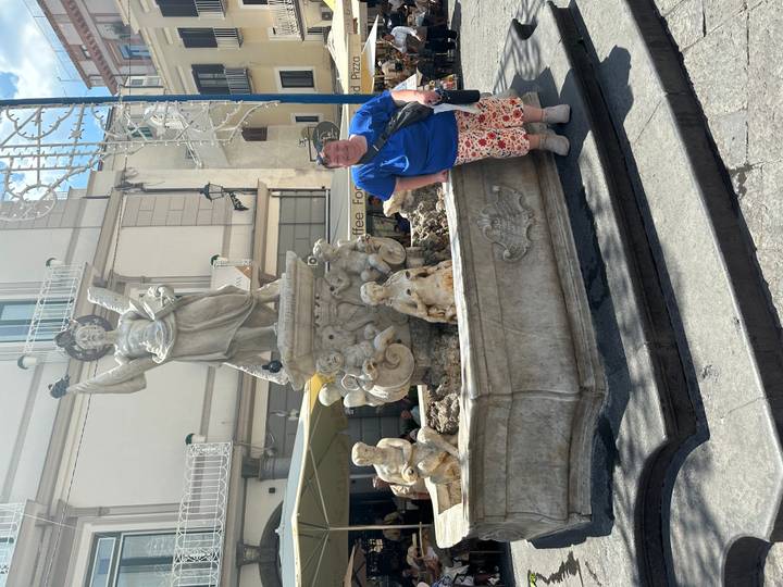 Woman leans on a stone fountain adorned with angel statue in a sunlit town square.