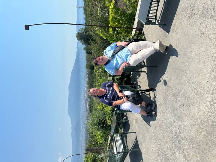 Two women seated on chairs at a hillside viewpoint overlooking countryside and distant mountains.