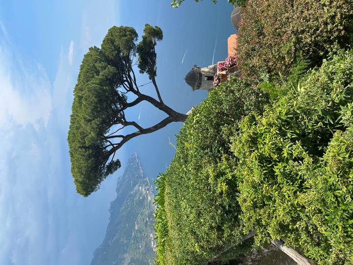 Panoramic view from a hilltop garden with a distinctive umbrella pine overlooking the deep–blue Amalfi Coast and a small domed stone tower.
