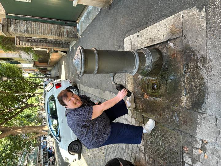 Visitor refilling a bottle at a traditional street fountain along a leafy urban lane with parked cars.
