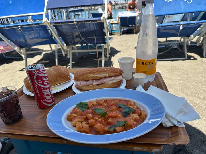 Beach-side meal setup with gnocchi, ham rolls, cola and sparkling water on a small wooden table in front of sun-loungers on sand.