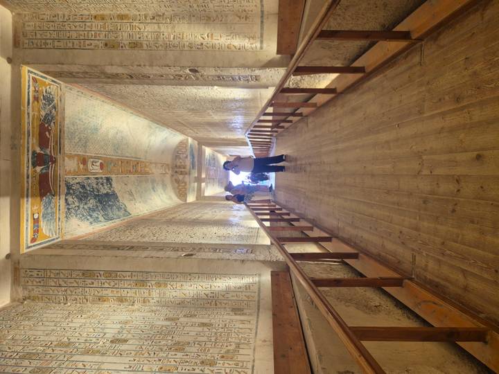 Decorated corridor with ancient hieroglyphs and colorful ceiling art inside a pharaonic tomb as visitors take photos.