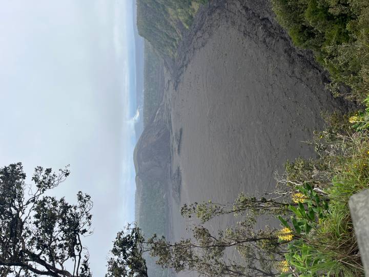 Wide view into a dark volcanic crater with steam rising in the distance under grey skies.