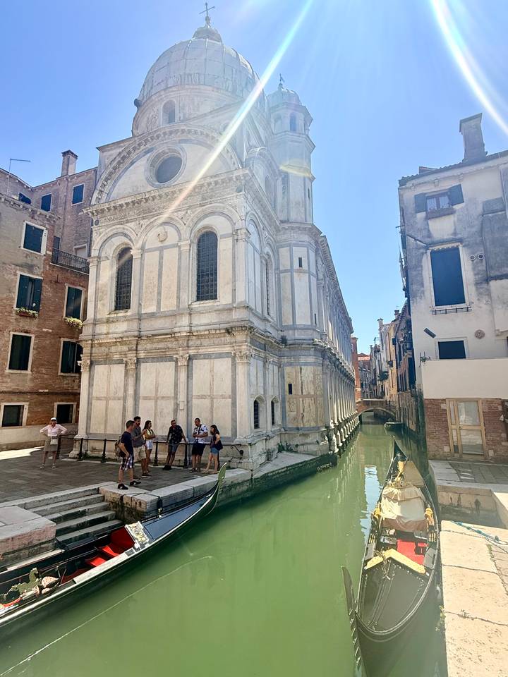 Travelers stand outside a marble-faced church beside a narrow Venetian canal.