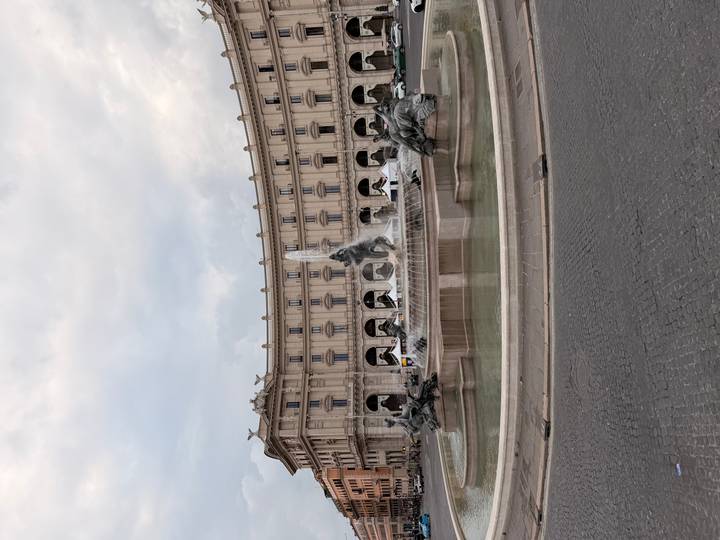 A grand fountain with bronze statues sits in a large Roman piazza backed by a curved neoclassical building.