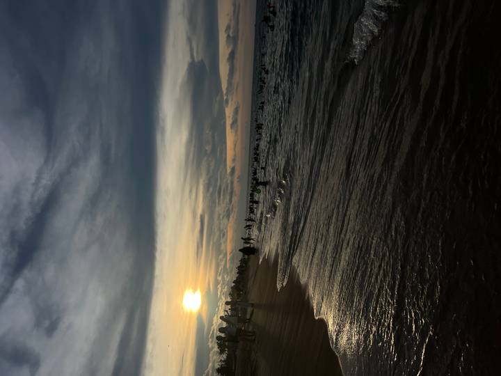Crowds swim and wade in the sea at dusk while the sun sets low over a sandy Vietnamese beach.
