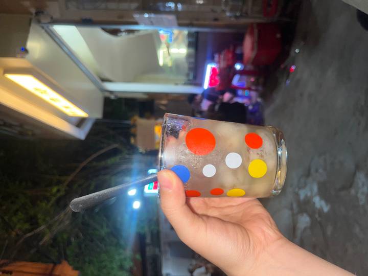 A hand holds up a polka-dotted glass of iced drink on a busy night-time street with small plastic stools in the background.