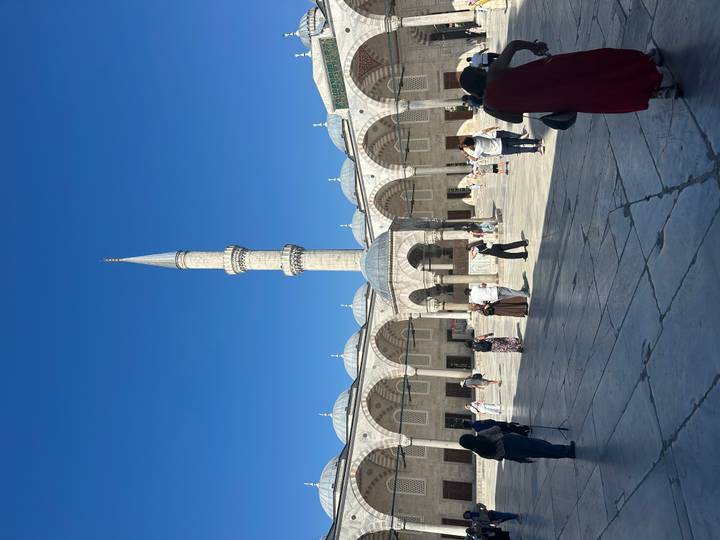 Visitors walk across the stone courtyard of an ornate Ottoman-era mosque dominated by a single tall minaret beneath a vivid blue sky.
