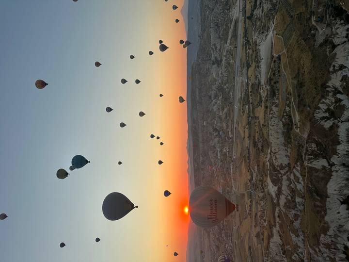 Dozens of hot-air balloons drift above the sculpted valleys of Cappadocia as the sun rises on the horizon, painting the sky orange.