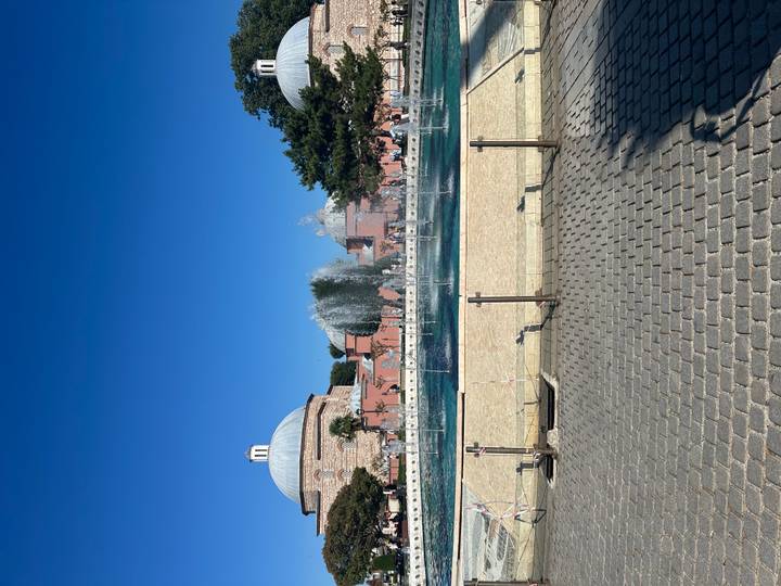 Public square with a large turquoise fountain pool framed by domed historic bathhouses and bright blue sky.