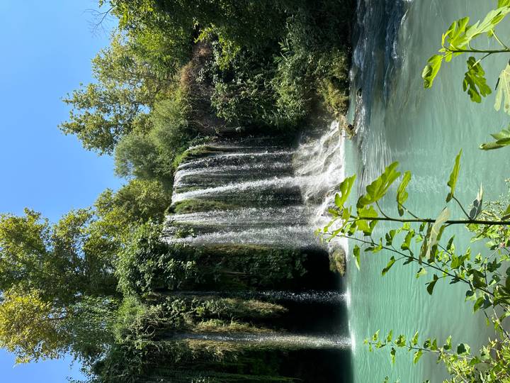 Crystal waterfall cascades into a turquoise pool surrounded by lush greenery under bright summer sunshine.