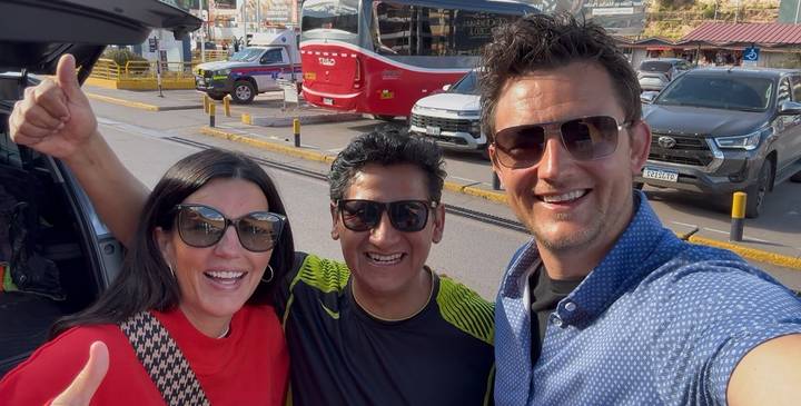 Three smiling travellers take a selfie outside a parked van at a Peruvian transport hub.