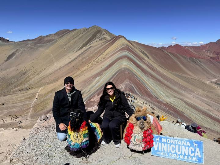 Two tourists pose with colorfully adorned llamas atop the striking rainbow hues of Vinicunca Mountain.
