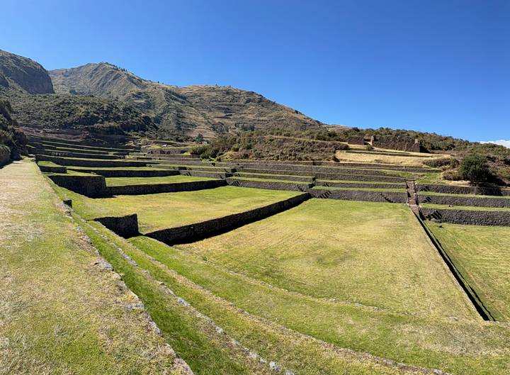 Terraced Inca ruins cascade down a green mountainside under a bright blue Andean sky.