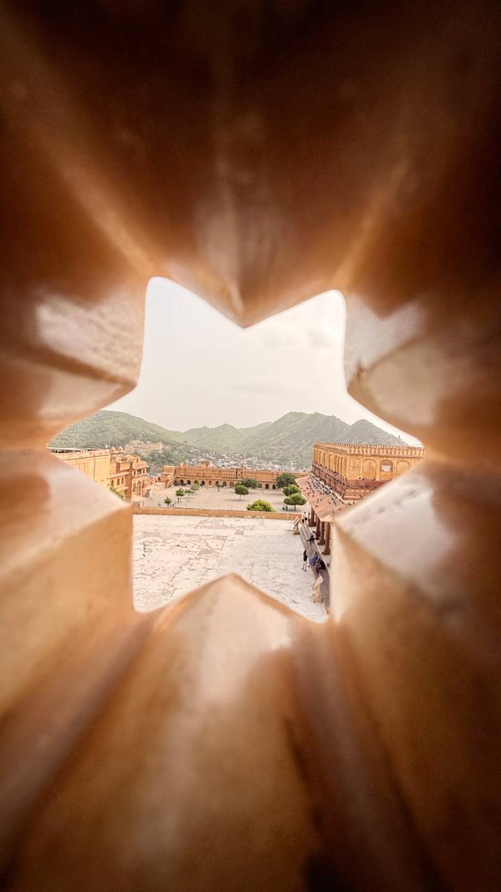 Creative peek through fortress lattice reveals Jaipur’s Amber Fort and surrounding green hills.