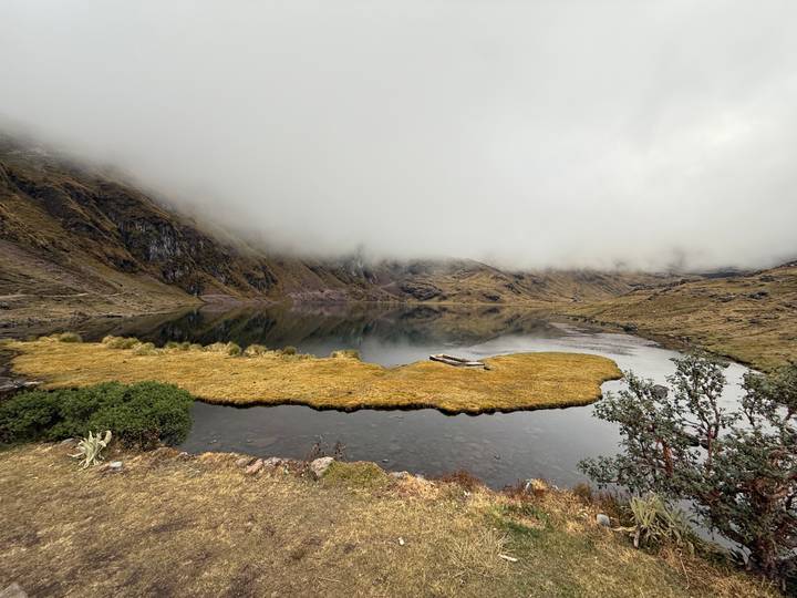 Serene high-altitude lake partly shrouded in mist with grass peninsula and mountains.