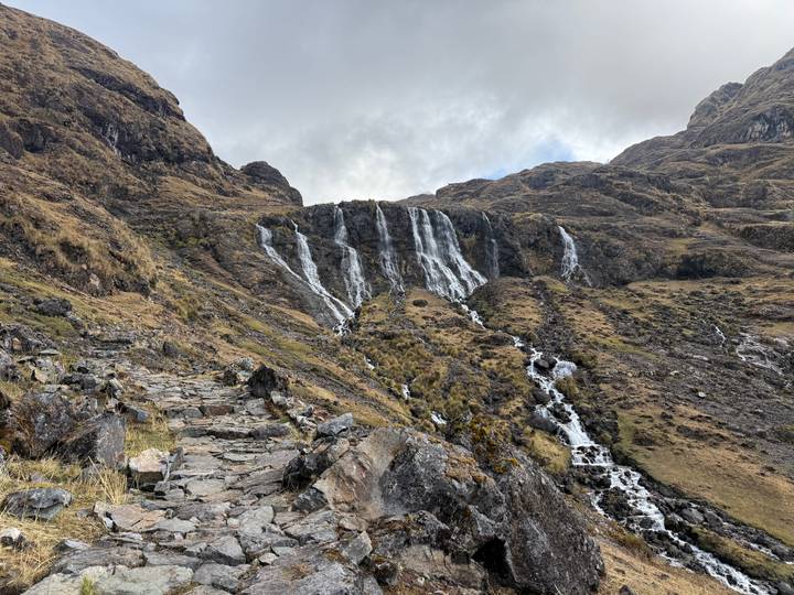 Multi-tiered waterfall cascading down rocky Andean mountainside beside stone trail.