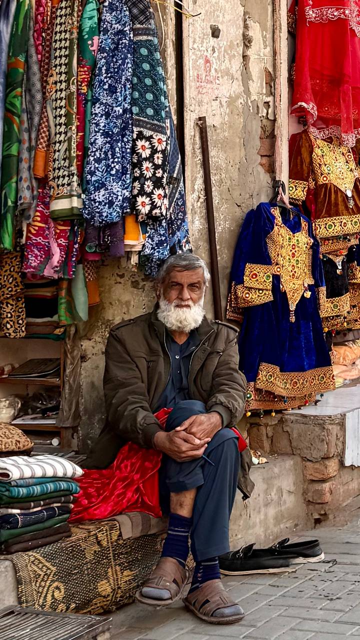 Portrait of elderly bearded man seated at colorful textile stall in Pakistan bazaar.