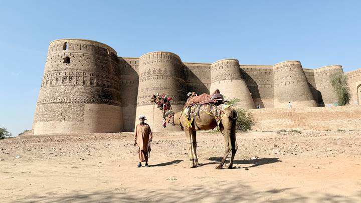 A man and a saddled camel stand on sandy ground in front of the massive brick bastions of an ancient desert fort under a clear blue sky.