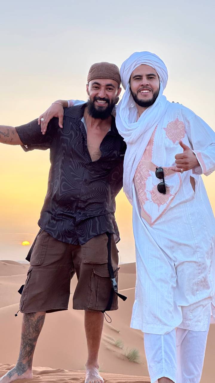 Close-crop of two friends in desert attire celebrating at sunset with golden sky behind.
