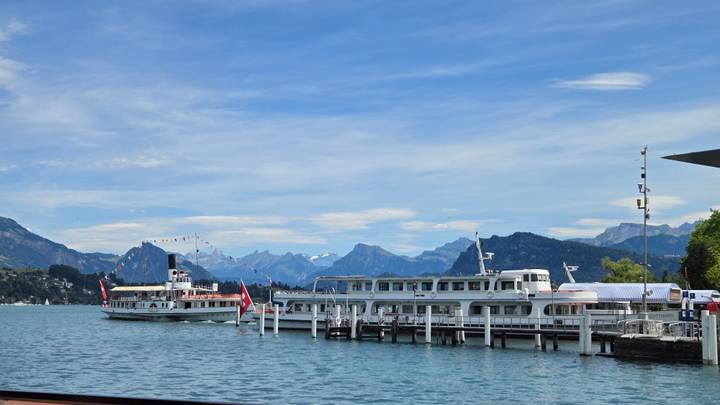 Boats moored on turquoise Lake Lucerne with distant snow-capped Alps under a bright blue sky.