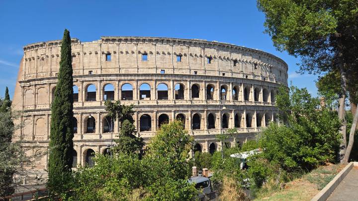 Panoramic view of the ancient Colosseum surrounded by green trees under a bright blue Roman sky.